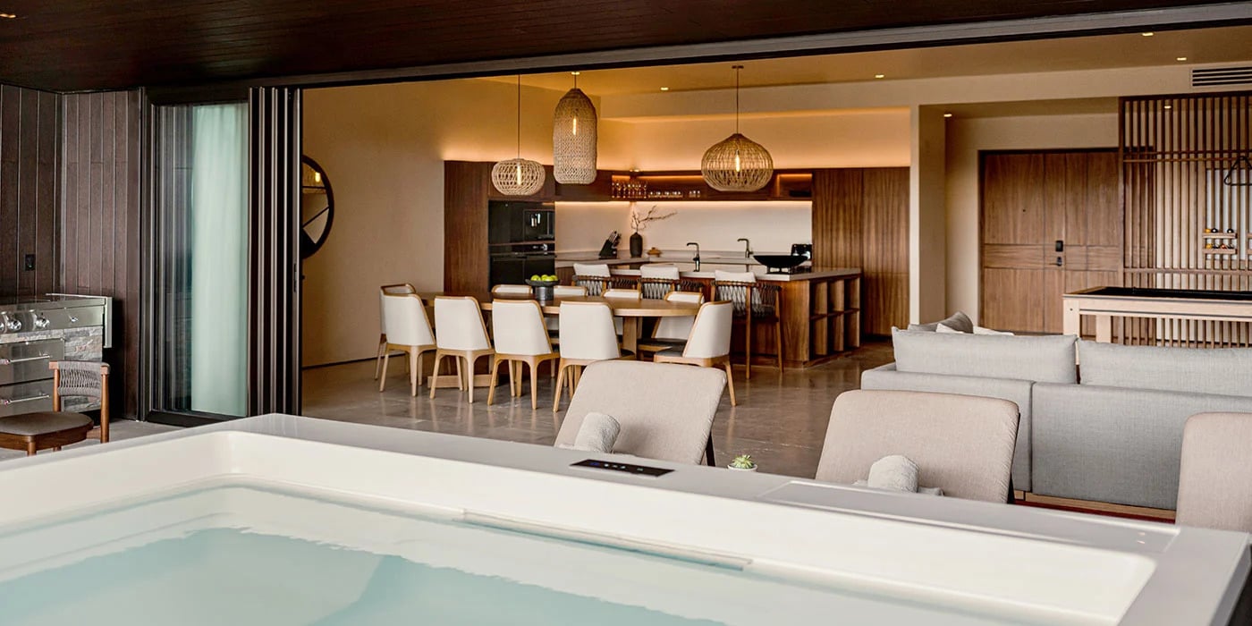 A modern, open-concept luxury residence featuring a jacuzzi in the foreground, with a seamless transition to an indoor dining and kitchen area. The space is decorated with warm wood tones, pendant lighting, and contemporary furnishings.