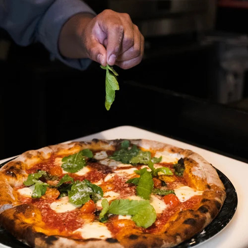 A chef’s hand is seen adding fresh greens to a wood-fired pizza with a crispy crust, melted cheese, and vibrant tomato sauce. The dimly lit setting enhances the gourmet dining atmosphere.