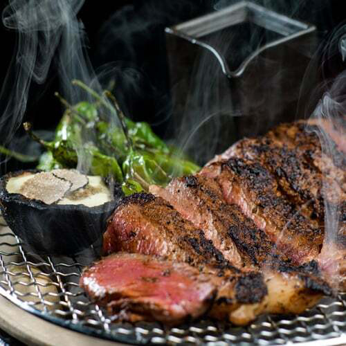 A perfectly grilled steak sliced into thick pieces, releasing steam, sits on a metal grill alongside blistered shishito peppers and a truffle-topped side dish. A glass box in the background adds a dramatic smoky effect.